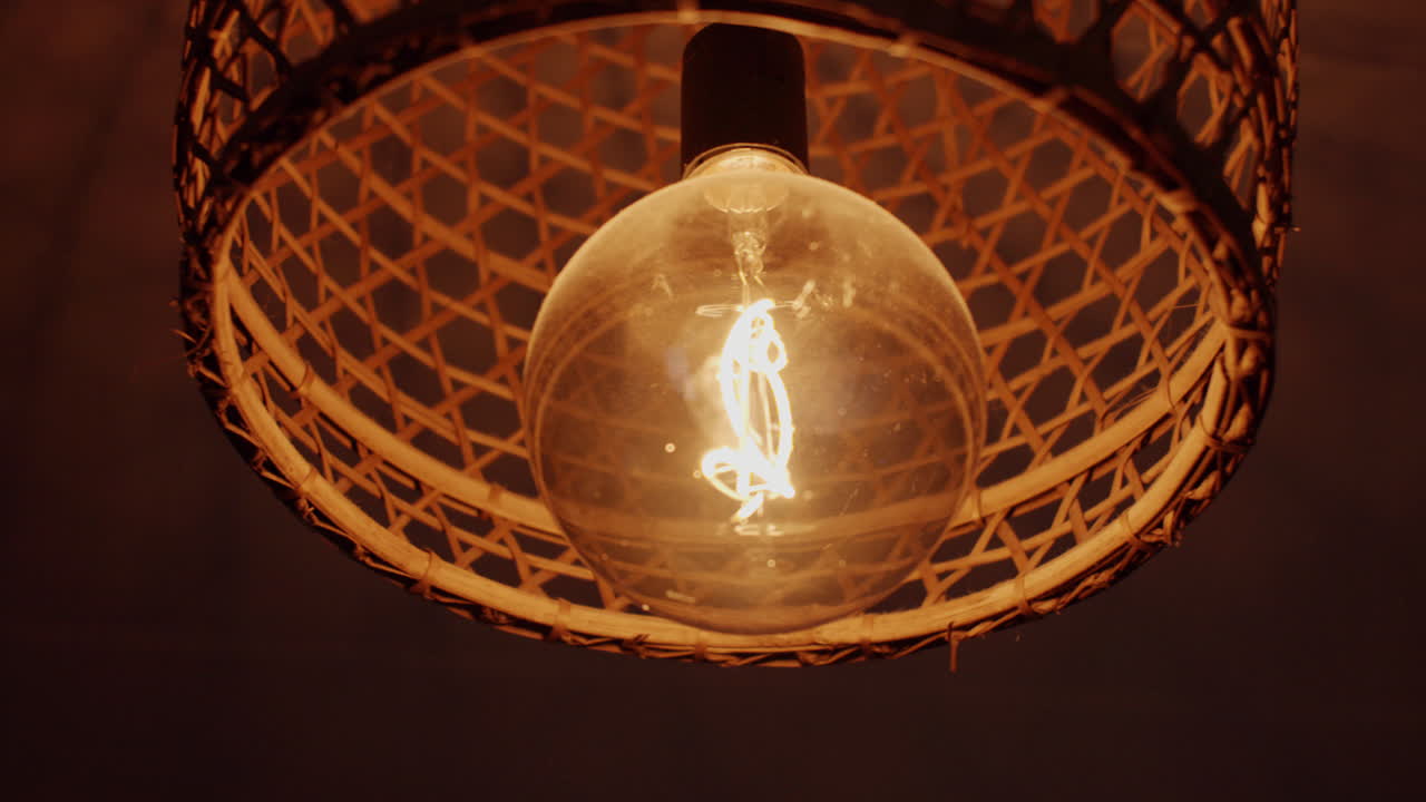Close-up footage of a glowing light bulb inside a woven wicker lampshade. The warm ambient glow creates a cozy atmosphere, highlighting texture and detail in a rustic, handcrafted interior design