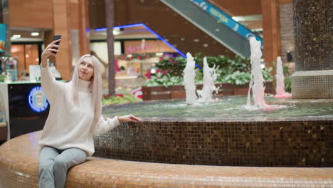 modern shopper sits by water fountain in busy mall scene, manicured hand skimming clear water while smiling and lifting smartphone for selfie, neon sign and escalator blurred in background