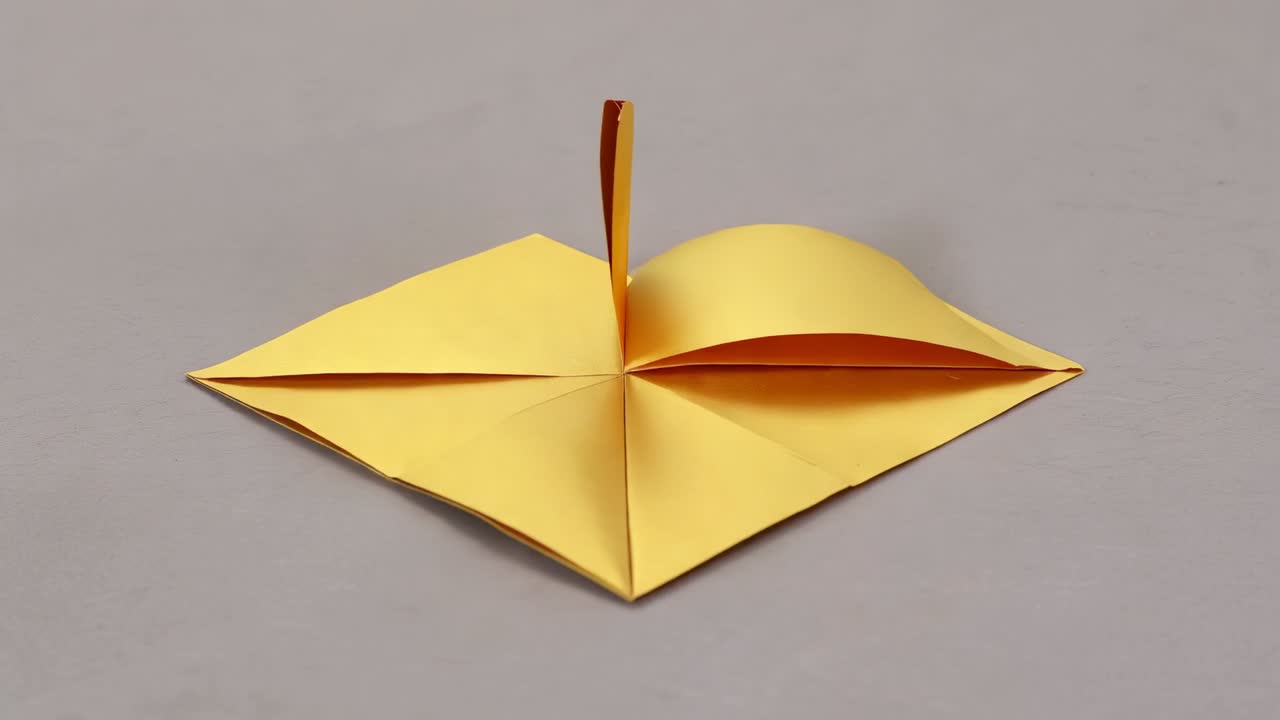 Starting crease lines guiding yellow square paper folding into diamond on gray mat, forming petals