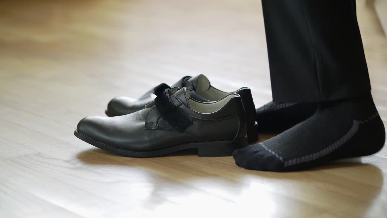 School boy is putting on black leather shoes on the wooden floor background. Men's shoes with velcro on the floor and a male in trousers puts them on at home. Close-up.