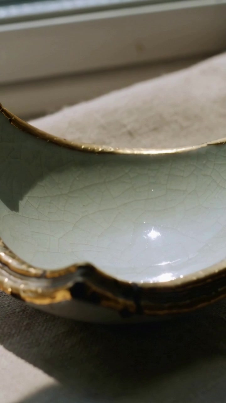Vertical video: Tilting camera showing ceramic dish on cloth at window, revealing gold rim, crackle