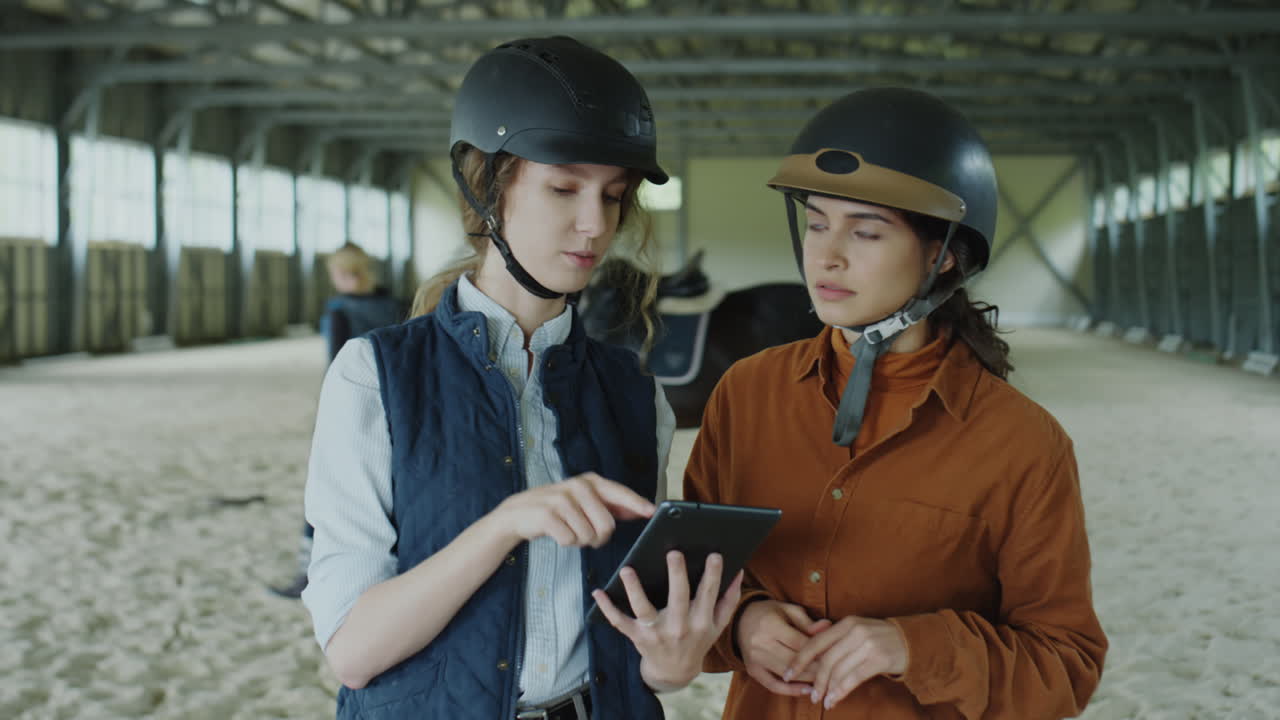 Two Women in Equestrian Gear Using a Tablet in an Indoor Horse Arena