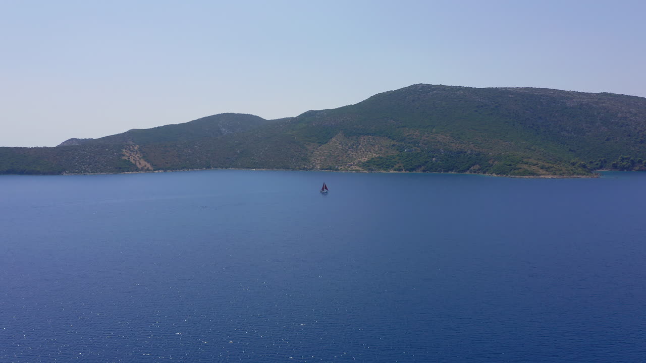 Aerial: A sailboat with red sails is sailing offshore on a clear summer day