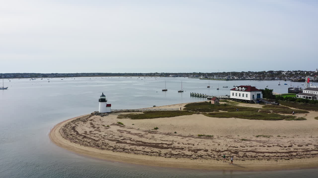 Drone orbita a sinistra intorno al Faro di Brant Point a Nantucket, Massachusetts, rivelando la costa sabbiosa, gli edifici portuali e le barche a vela ancorate in un'ampia ripresa aerea costiera