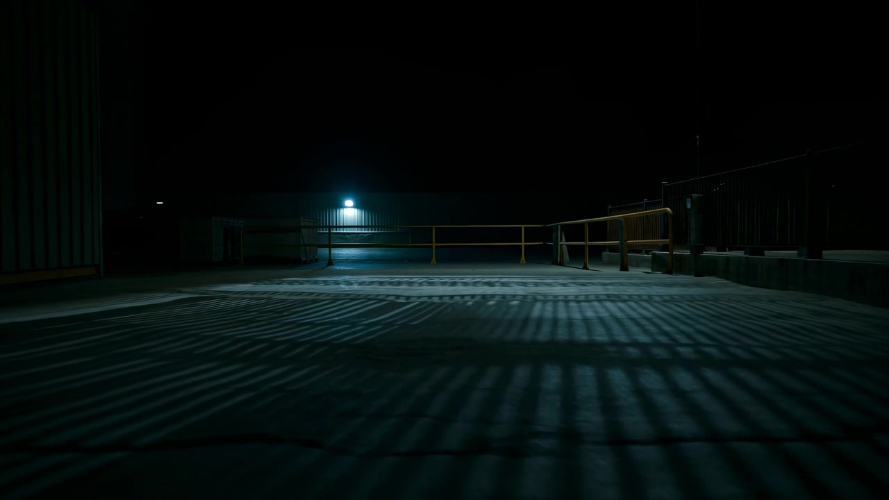 Wide low angle night view of empty industrial rooftop or loading area, lit by single cool white wall lamp that casts pronounced parallel shadow stripes across concrete, metal railings, corrugated