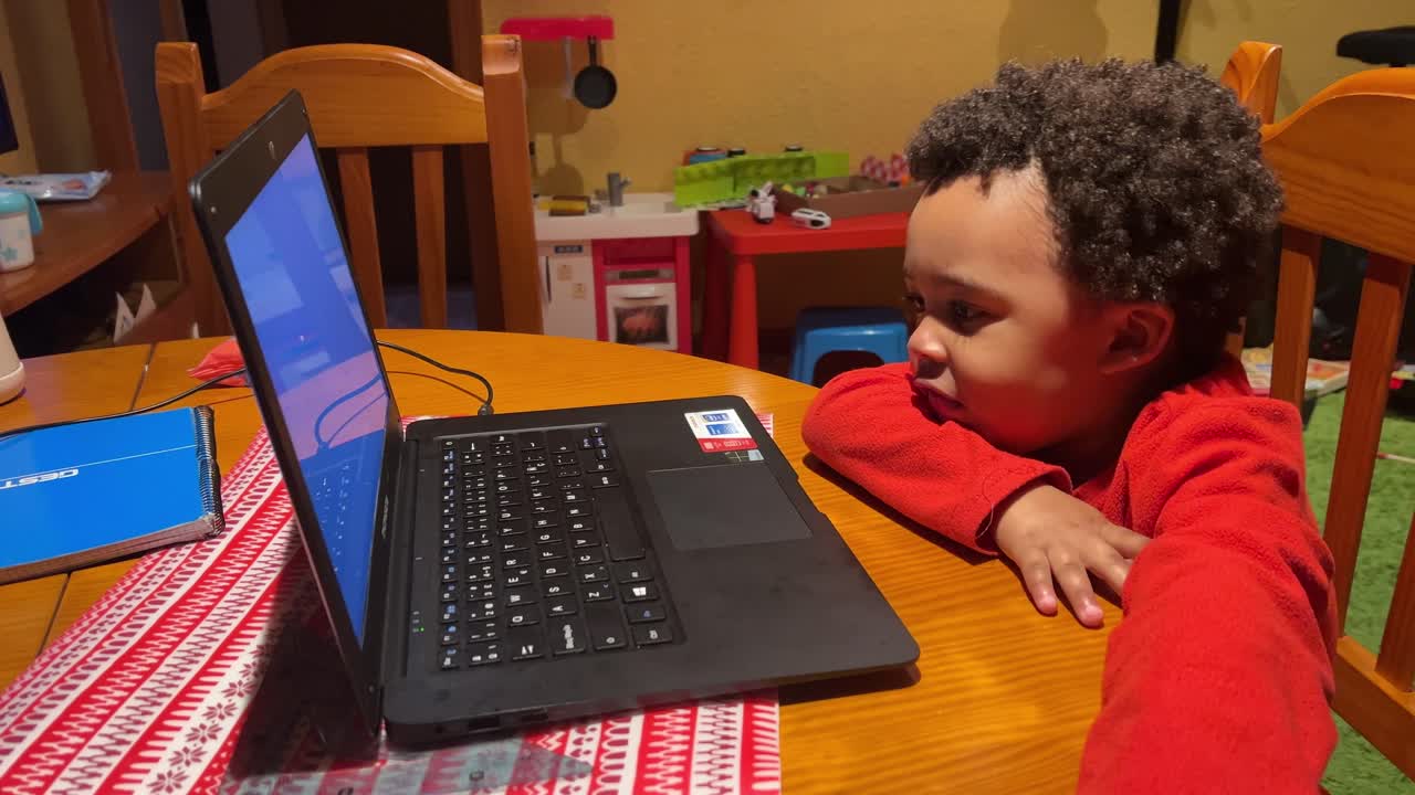un gracioso bebé negro de dos años juega con una laptop en casa, sentado en una silla de madera