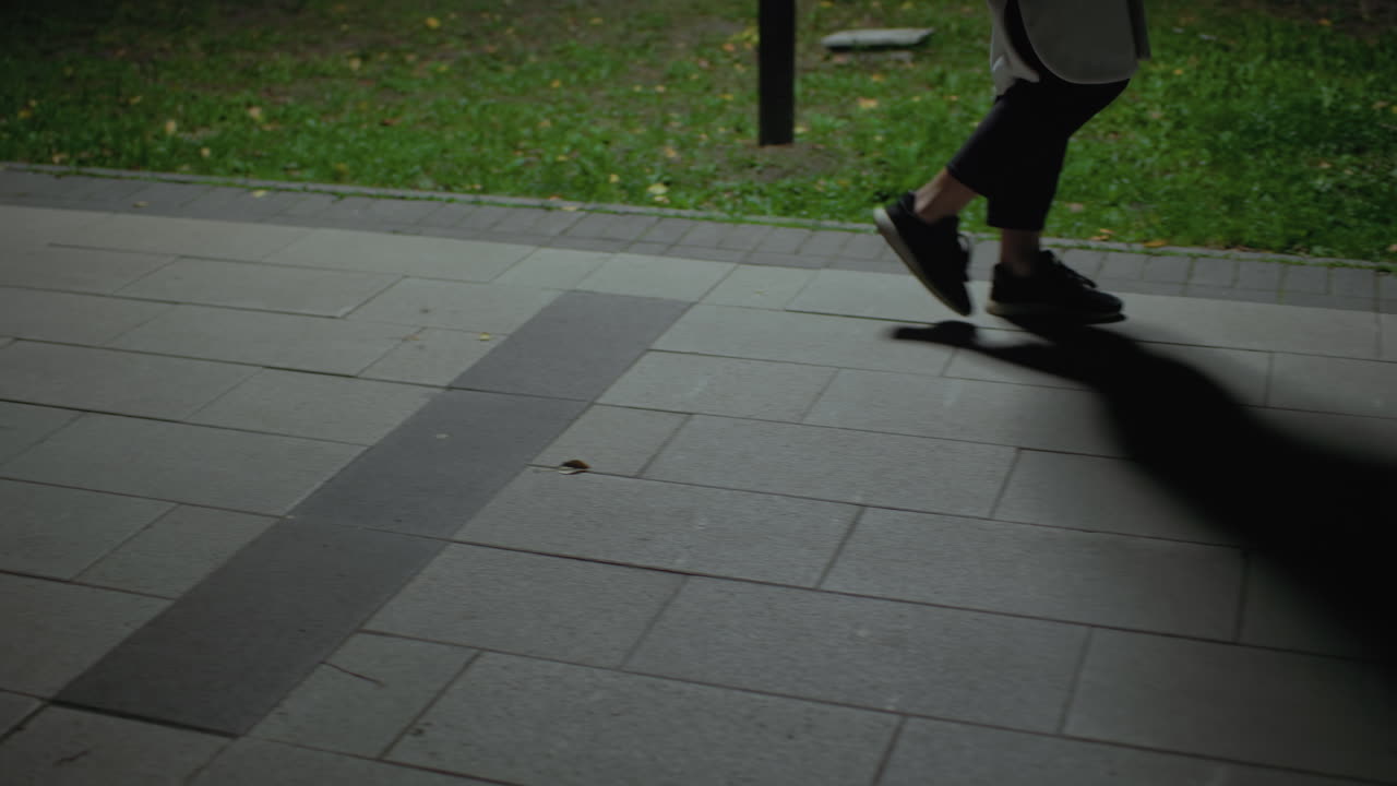 Leg view person walking along paved surface near grass, dim environment casting distinct shadows across walkway, capturing subtle motion in quiet night setting, footwear and gentle light