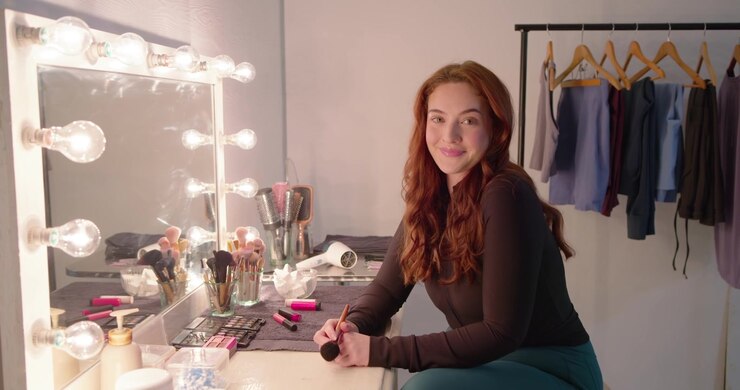 Woman in 20s preparing for shoot, leaning at lighted vanity and applying face makeup with brushes