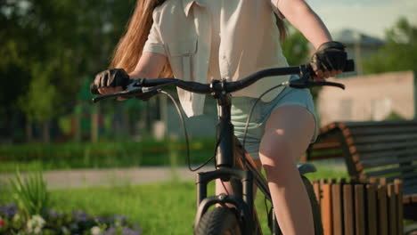 close-up of cyclist preparing for outdoor ride, mounting bicycle near vibrant garden with colorful flower beds, surrounded by trees and wooden bench, with blurred figure in background