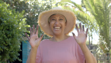 portrait of happy senior biracial woman with dirty hands wearing hat in garden at home, slow motion