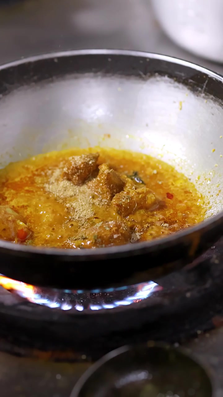 Primo piano di una mano che aggiunge spezie a un curry giallo che sobbolle in una cucina professionale.