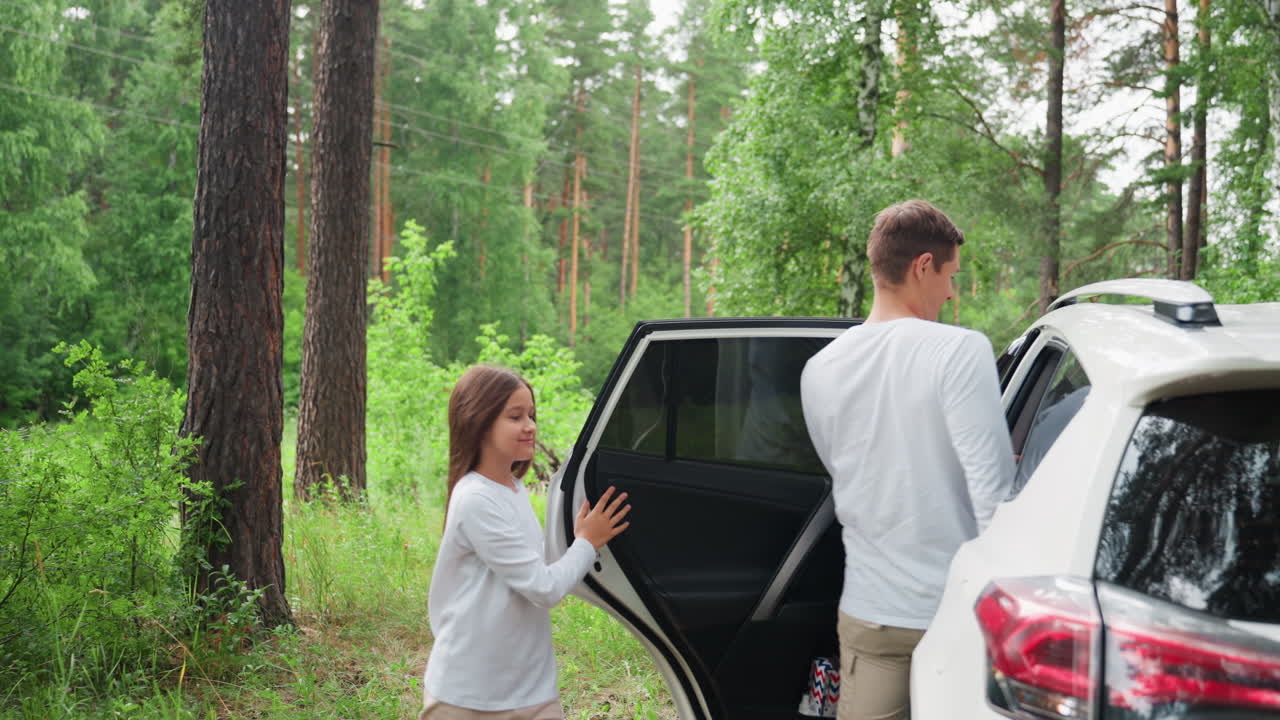 Dei giovani salgono su un'auto bianca parcheggiata in una tranquilla foresta verde, circondata da alberi e natura incontaminata, godendosi un momento di calma all'aria aperta durante un viaggio in famiglia con la luce del sole che filtra tra le foglie.