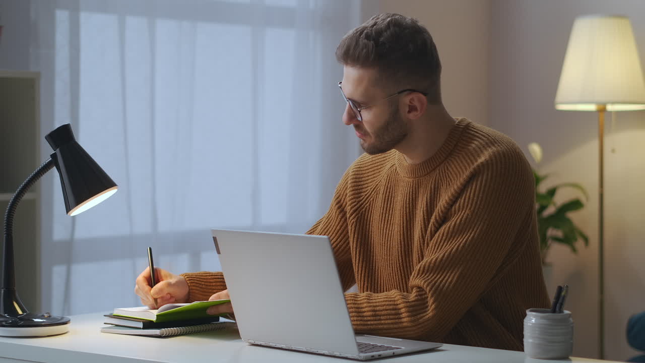 adult man is writing notes in notebook and typing at laptop sitting alone in room working remotely using internet connection for work medium portrait