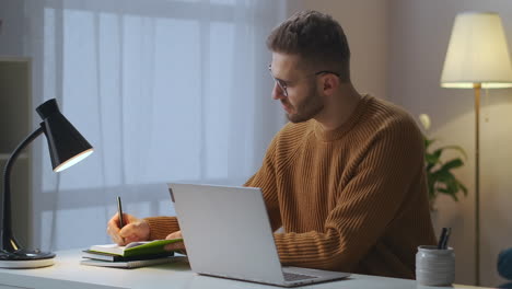 Ein-Erwachsener-Mann-Macht-Sich-Notizen-In-Ein-Notizbuch-Und-Tippt-Am-Laptop,-Sitzt-Allein-Im-Zimmer-Und-Arbeitet-Aus-Der-Ferne-über-Eine-Internetverbindung-Für-Die-Arbeit,-Mittleres-Porträt