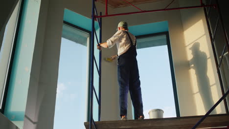 painter primes the wall, stands high on the scaffolding inside the house