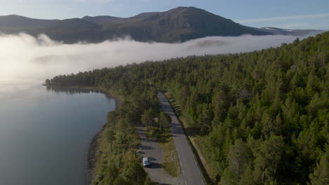 aerial drone view flight over scenic green pine tree forest and country road with mountain range in background and mystical cloudy fog