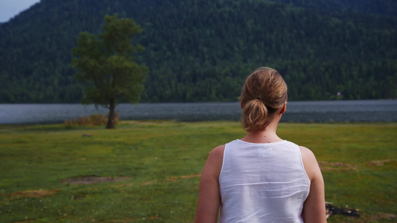 Woman stands in grassy field near lake. Mountains loom behind calm water. She gazes peacefully toward distant trees. Serene moment perfect for travel or nature content. Could use in wellness