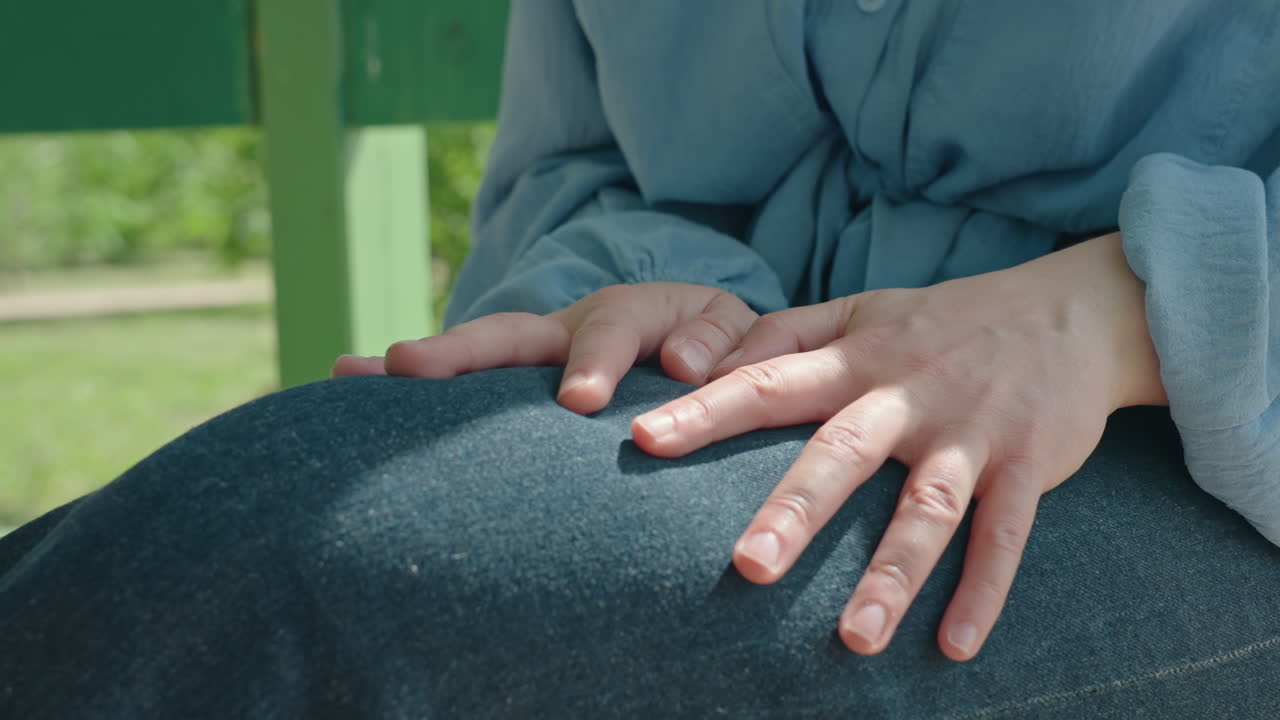 Primer plano de una mujer caucásica con las manos entrelazadas y apretadas sobre el regazo de tela vaquera, con los nudillos enrojecidos por el sol, tensión emocional mezclada con esperanza; las mangas de la camisa azul y el banco del parque crean un íntimo momento espontáneo.
