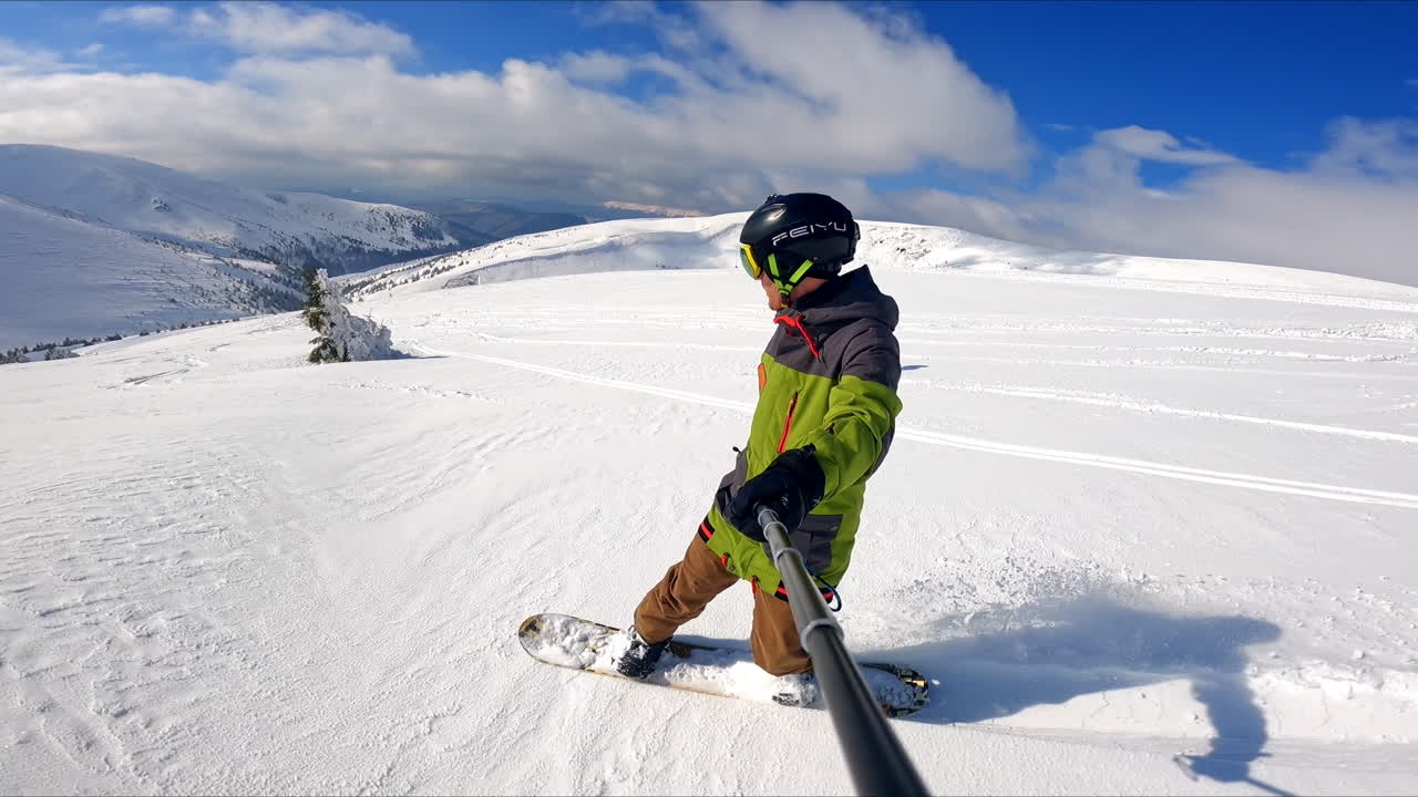 Modernly equipped snowboarder taking selfie of the ride. Few sportsmen go down the slope at backdrop.