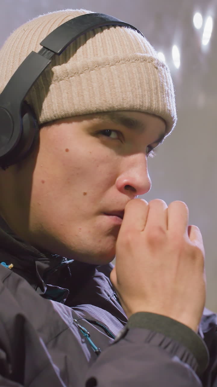 후디를 입고 헤드폰을 착용한 젊은 성인이 담배를 피우는 동안 금속 벽에 앉아 차분하고 상적인 표정을 보이고 있습니다.