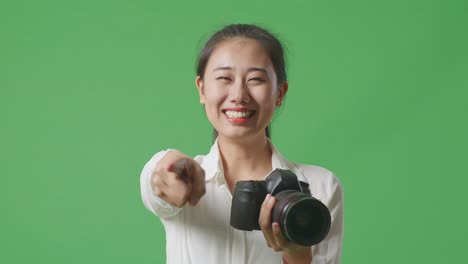 close up of asian photographer using a camera taking pictures and smiling touching her chest while standing on green screen background in the studio