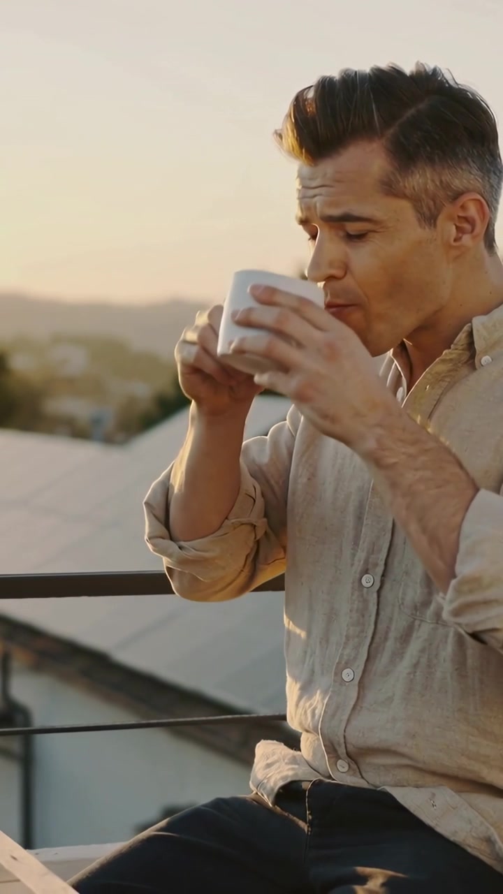 Video vertical: Hombre bebiendo con camisa de lino, levantando una taza blanca en un tejado al atardecer, relajándose, con espacio para texto.