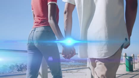 Animation-of-light-moving-over-midsection-of-couple-walking-by-beach-holding-hands