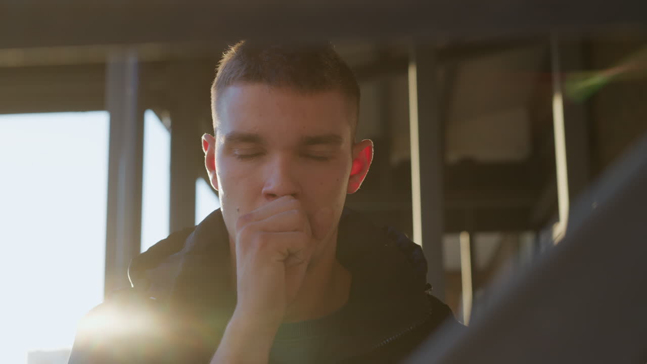 close up of sick student coughing outdoors with hand over mouth, illuminated by strong glow of sunlight from behind, standing in casual jacket near urban metal structure during golden hour