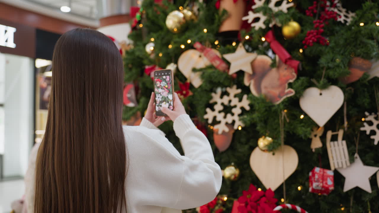 Back view of woman taking picture of tall Christmas tree adorned with ornaments and lights in a vibrant mall, bright lights filter through the mall modern architecture