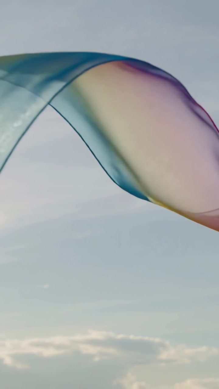 Video vertical: Cinta suave y transparente deslizándose y formando bucles a través de un cielo pálido con nubes bajas, movida por el viento.