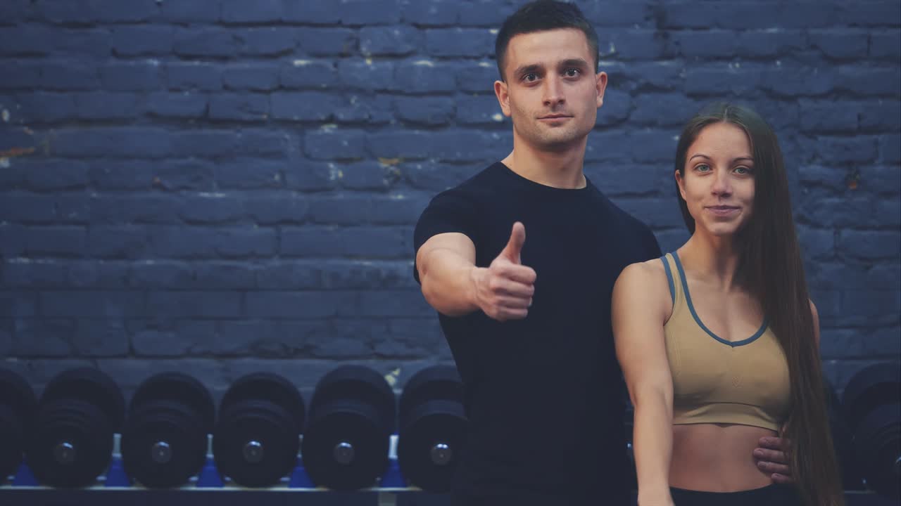 una hermosa joven y su bien construido novio juntos muestran una clase de gestos. se alegran de verse el uno al otro en el gimnasio. los jóvenes están listos para comenzar sus entrenamientos.
