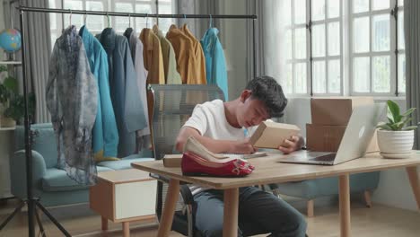 Asian Man Online Seller Looking At The Package And Writing In The Notebook While Using Computer For Selling Clothes At Home
