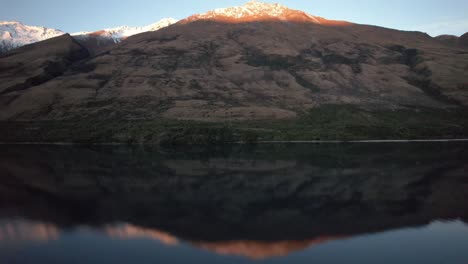 Vista-Aérea-Volando-Hacia-Atrás-Para-Revelar-El-Perfecto-Reflejo-De-La-Montaña-Al-Atardecer-Con-La-Montaña-Cubierta-De-Nieve-Atrapando-El-Alpenglow-En-Queenstown,-Nueva-Zelanda