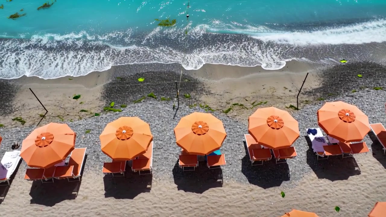 Drone in volo che riprende due adulti che passeggiano tra ombrelloni arancioni sulla spiaggia, mostrando la marea