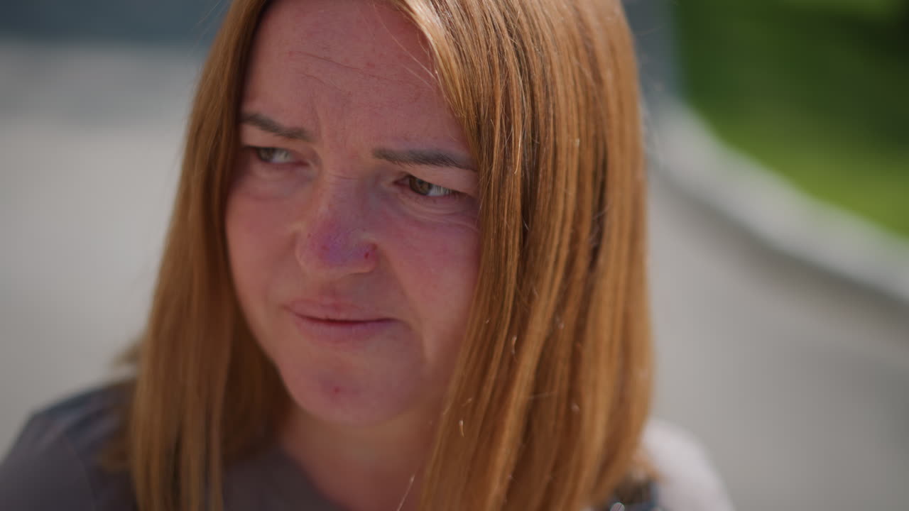 Close up of uneasy lady looking unsettled outdoors with blurred green background, sunlight highlighting hair and face, expression conveying anxiety, reflection, emotional tension