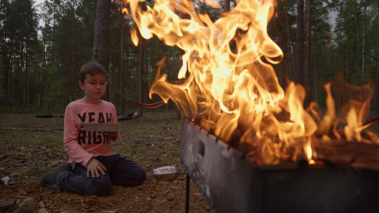 바베큐 그릴에서 밝은 불꽃은 불 근처에 앉아있는 소년의 실루 ⁇ 을 조명