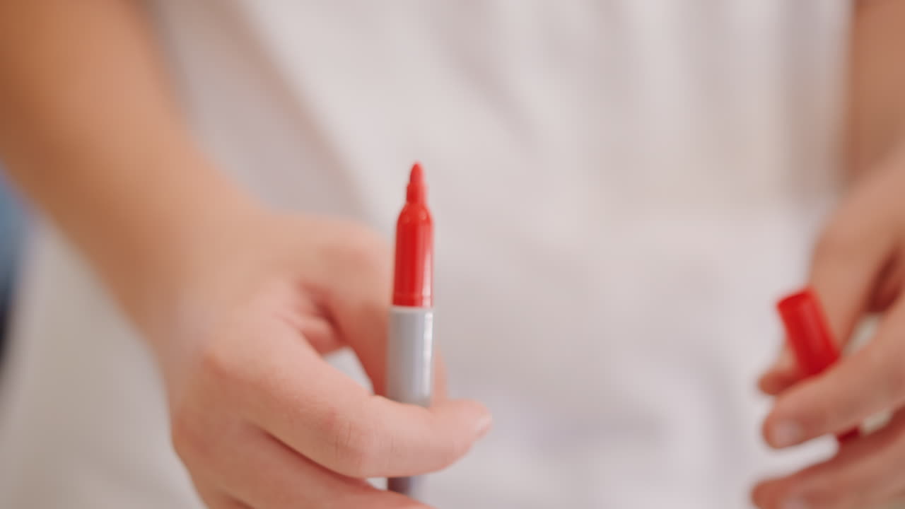 Primer plano de la eliminación de la tapa de un rotulador rojo, manos preparándose para dibujar, un niño pequeño quitando la tapa de un rotulador de fieltro con agarre suave, fondo de camisa blanca, iluminación cálida y suave, poca profundidad de campo, sereno