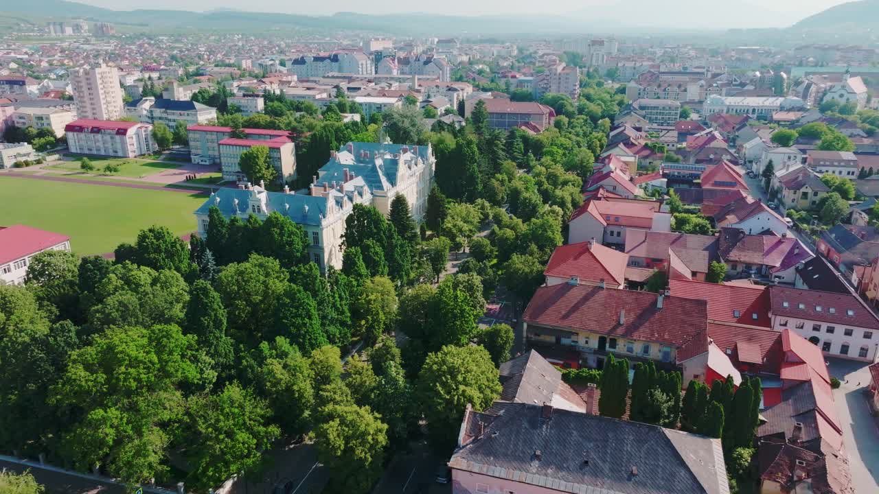 A panoramic aerial photo captures an impressive historic building set in a green park, a large sports field, and buildings against the backdrop of a sunny European city. Bistrita, Romania