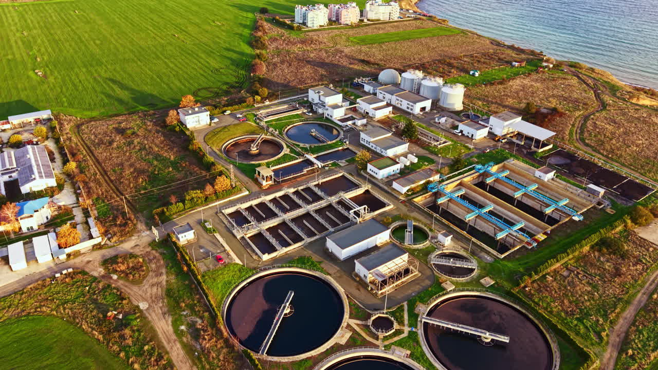 Water treatment facility near the coast surrounded by green fields and urban buildings, bustling with activity during sunny daylight hours