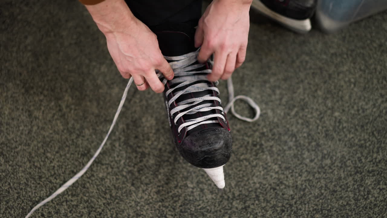 Close-up view of hands loosening the laces of a black ice skate with red accents, on a gray carpeted floor, the person is wearing black trousers