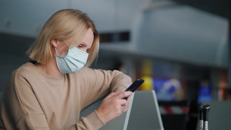 A-woman-in-a-protective-mask-is-waiting-for-her-flight-in-the-airport-terminal,-using-a-smartphone