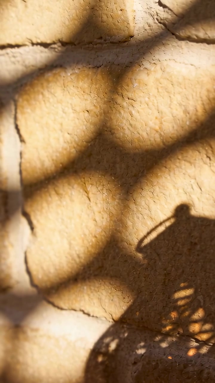 Vertical video: Shifting sun casting scalloped shadows on stucco wall at patio tilting woven shadow