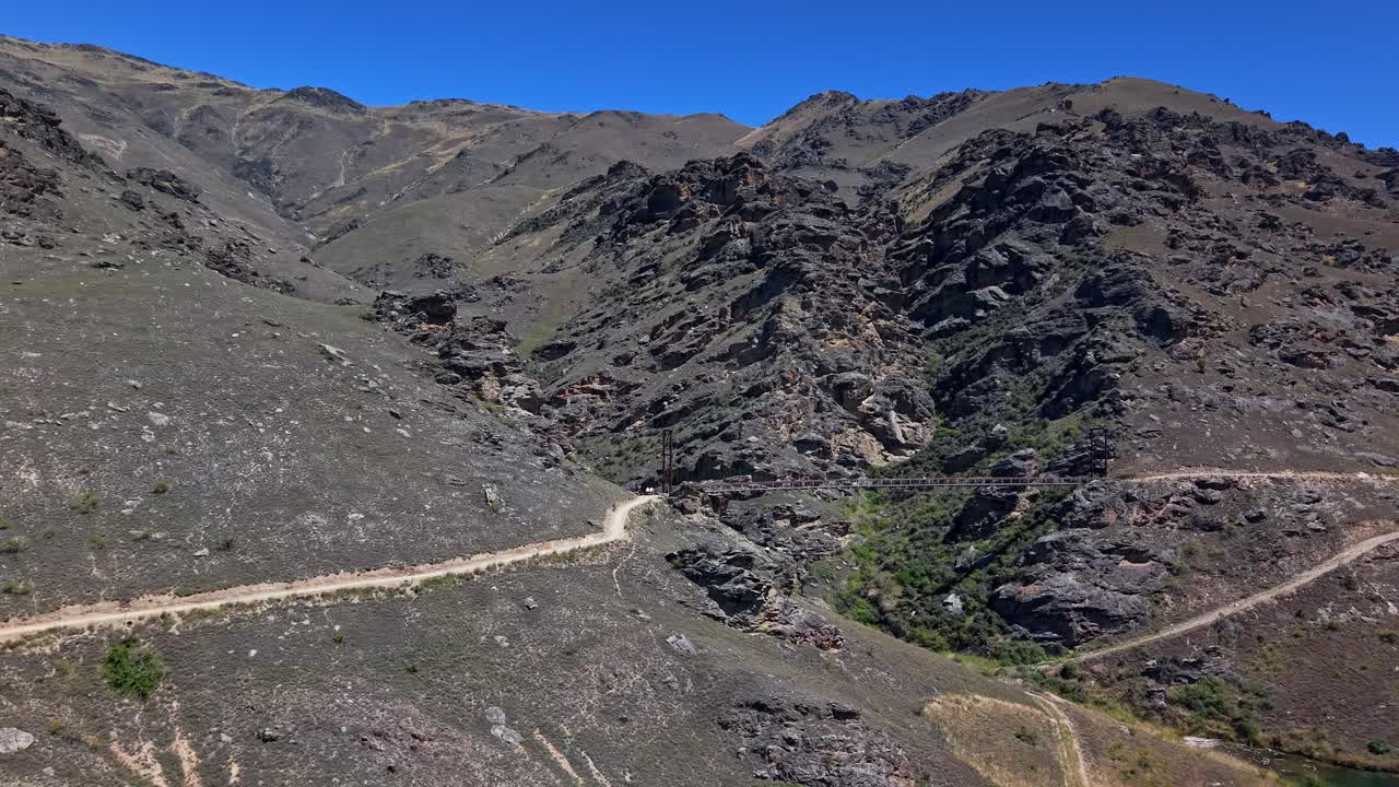 Thrilling drone footage of cyclist biking across 86m high bridge suspended over rugged rocky gully on scenic cycling adventure outdoors in fresh air and summer sunshine in Cromwell, New Zealand
