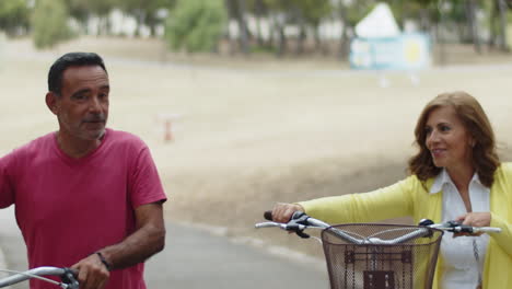 Front-view-of-happy-mature-couple-walking-with-bikes-in-park