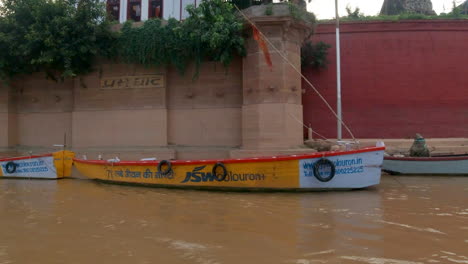 Cinematic-Varanasi-India-Ganges-River-cruise-canal-boats-person-Northern-State-large-gathering-at-shore-Ancient-Holy-city-Ghat-Pradesh-Province-landscape-gray-cloudy-afternoon-muddy-brown-follow-left