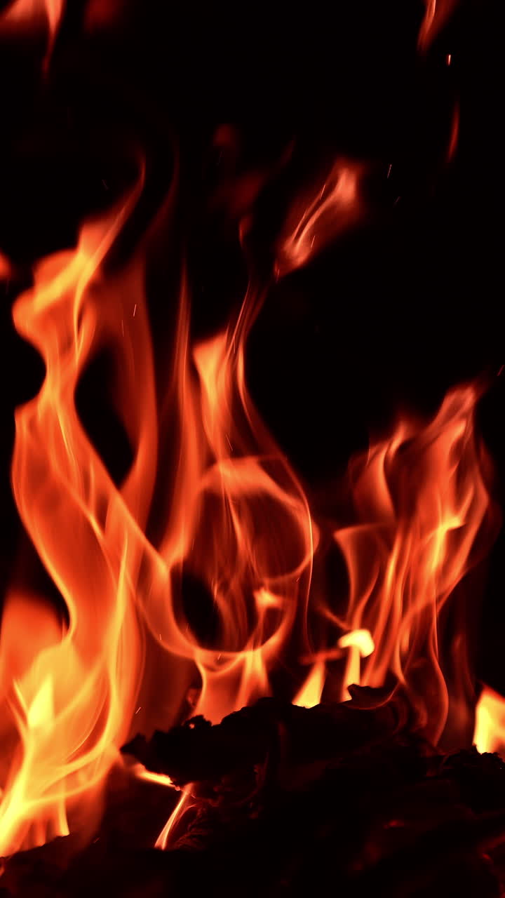 Wooden logs burning in the campfire at night. One sticks breaks and multiple sparkles rise up. Vertical video