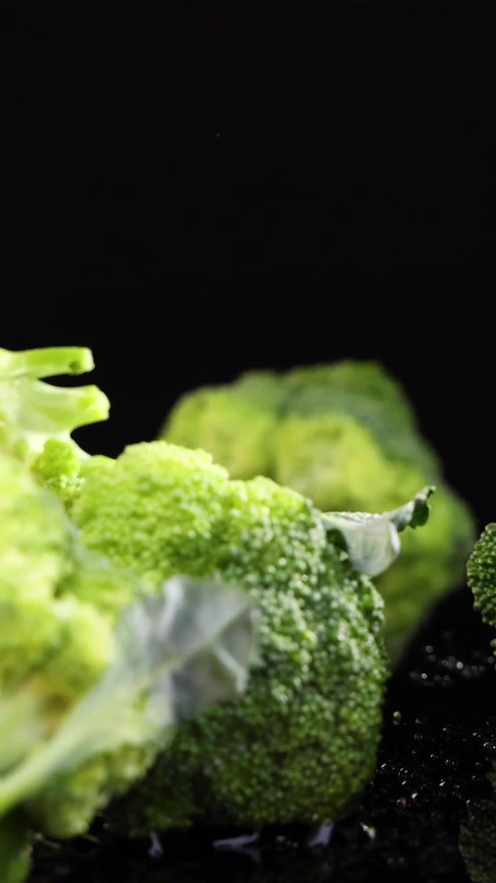 Cimette di broccoli cadono e rimbalzano su una superficie nera riflettente, con goccioline d'acqua che schizzano, sotto una brillante illuminazione da studio con effetto slow-motion e telecamera fissa