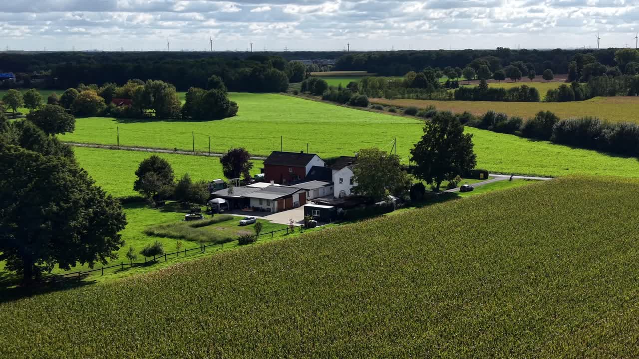 Farmstead house surrounded by green fields and riding cyclist on street. Sunny day with clouds at sky in summer. Aerial orbit wide shot. Modern wind turbines in distance