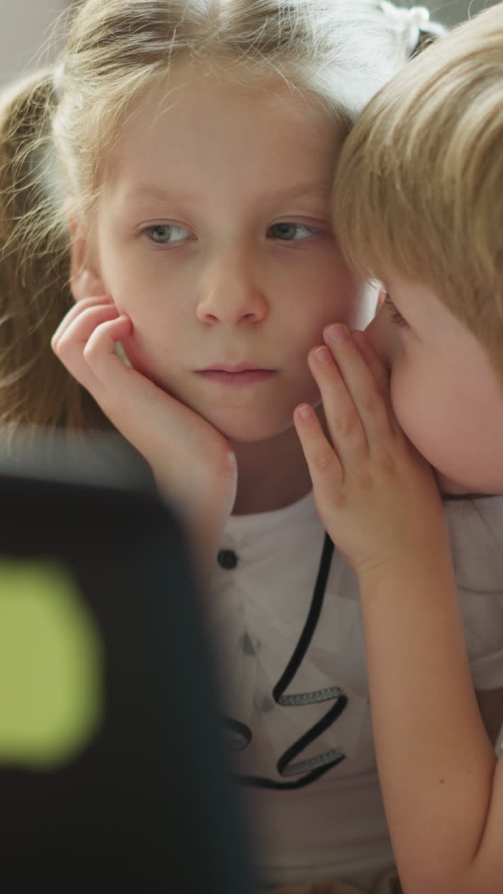 남동생은 노트북을 들고 테이블에 앉아 있는 여동생에게 비밀을 말하고, 소녀는 귀를 기울이고 머리를 기울이고, 친절한 형제자매는 집에서 함께 시간을 보낸다.