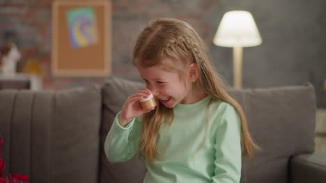 Joyful-little-girl-shakes-bottle-with-golden-glitter-on-sofa